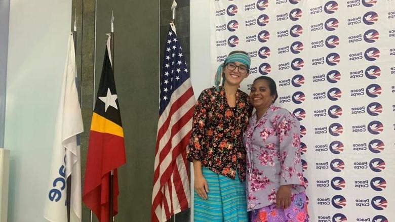 Two people smile, arm-in-arm, wearing colorful garb in front of a Peace Corps banner